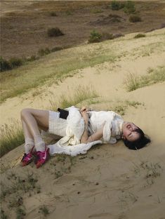 a woman laying on top of a sandy beach next to grass and sand dunes in the background