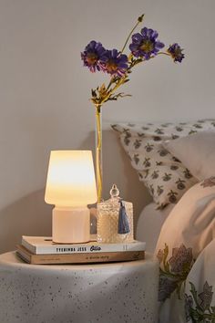 a table with a lamp and flowers on top of it next to a bed in a bedroom