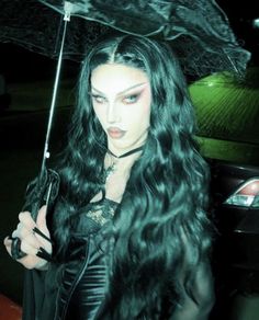 a woman with long black hair and makeup holding an open umbrella in her hand while standing next to a car