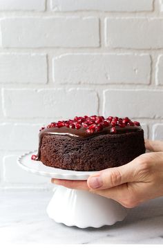 a person holding a chocolate cake with pomegranates on top, in front of a brick wall