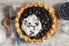 a pie with blueberries and whipped cream on top sitting on a wooden table next to a glass of water