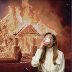 a woman is drinking from a mug in front of a house that was on fire