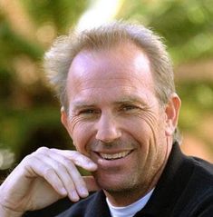 a man in a black shirt and tie smiles at the camera with his hand on his chin