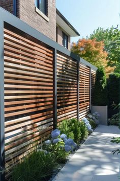 a wooden fence in front of a building with blue flowers and bushes on the side