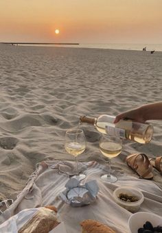 a person is on the beach with food and wine in front of them as the sun sets