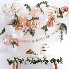 a table topped with cake and balloons next to a wall covered in greenery, foliage