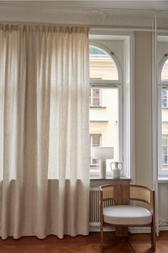 a chair sitting in front of a window next to a white curtain and table with a lamp on it