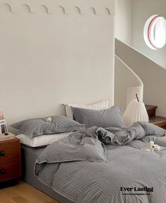 an unmade bed with grey sheets and pillows next to a wooden dresser in a white walled room