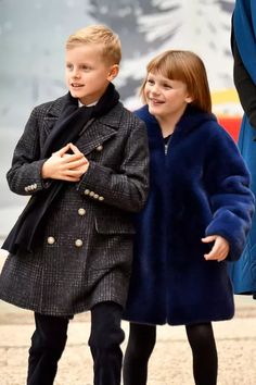 two children are walking down the street in coats and black pants, one is wearing a blue coat