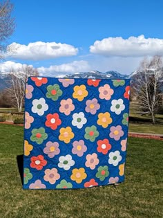 a blue quilt with colorful flowers on it in the middle of a grassy field near mountains