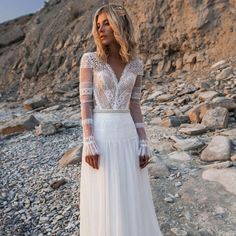 a woman standing on rocks wearing a white dress with sheer sleeves and beaded detailing