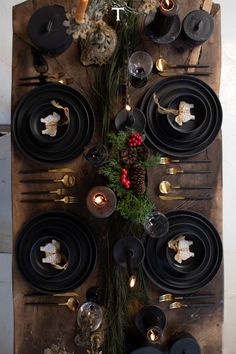 the table is set with black plates and silverware, pine cones, evergreen branches, candles, and christmas decorations