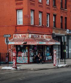 an old red brick building on the corner of a street with people sitting outside it