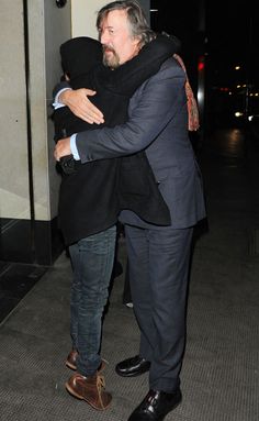 two men hugging each other in front of a building at night time, one is wearing a suit and the other has his head wrapped around his neck