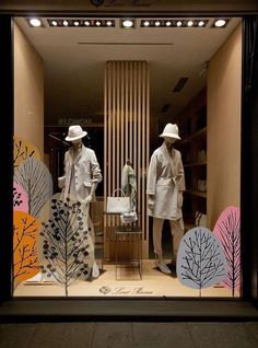 two mannequins dressed in white standing behind a window display with trees and leaves