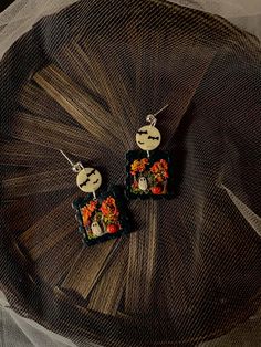 two square earrings with flowers on them sitting on top of a netted tablecloth