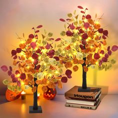 two fake trees with lights in them sitting on a table next to books and pumpkins