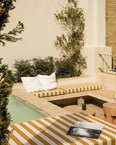 an outdoor lounge area with striped cushions and tables next to a swimming pool in front of a brick building