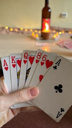 a hand holding four playing cards in front of a table with candles and bottles on it