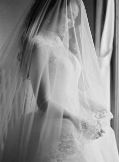 black and white photograph of a bride in her wedding dress looking out the window with her veil over her shoulder