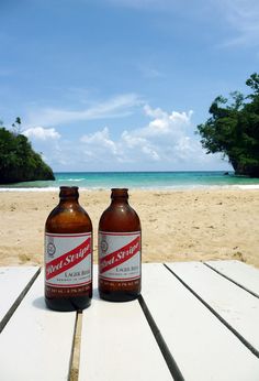 two beer bottles sitting on top of a wooden table next to the ocean and beach