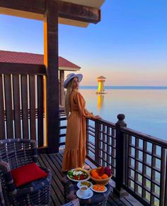 a woman standing on a balcony next to a table full of food and drinks with the ocean in the background