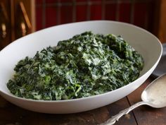 a white bowl filled with spinach on top of a wooden table next to a spoon