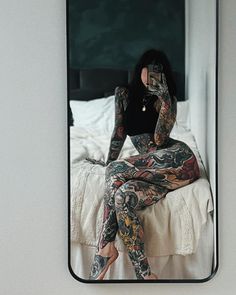 a woman with tattoos taking a selfie in a mirror while sitting on a bed