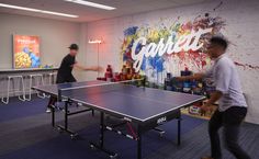 two men playing ping pong in an office setting with colorful graffiti on the wall