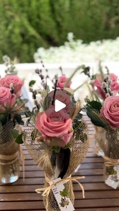 several vases with pink roses and greenery are on a wooden table in front of an outdoor setting