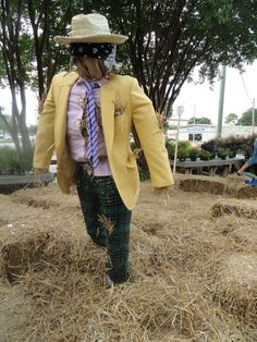 a scarecrow wearing a yellow jacket and tie walking through hay bales with trees in the background