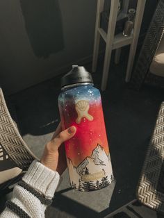 a person holding up a water bottle with mountains and stars painted on the side, in front of a chair