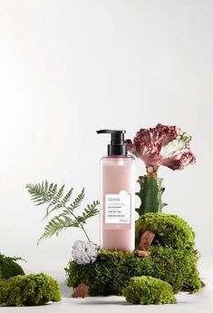 a bottle of lotion sitting on top of moss covered ground next to flowers and plants