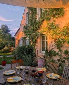 an outdoor dining table with plates and glasses on it, in front of a house