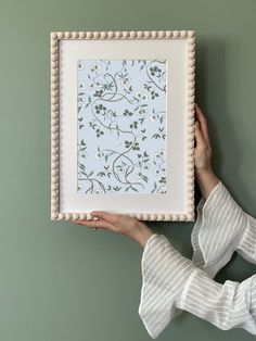 a woman is holding up a framed painting with flowers on it's side, against a green wall