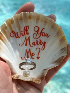 a person holding a shell with a ring on it that says will you marry me?