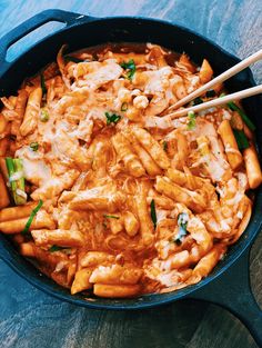 a skillet filled with pasta and vegetables on top of a wooden table next to chopsticks