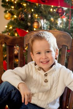 a young boy sitting in front of a christmas tree wearing a white sweater and jeans