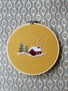 a small red house with snow on the ground and trees in front of it is embroidered onto a yellow hoop