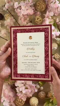 two hands holding up a red and white wedding card in front of pink flowers on a table