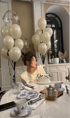 a woman sitting at a table with balloons and cake in front of her, surrounded by tea cups and saucers