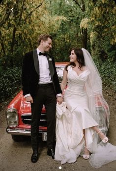 a bride and groom standing next to an old red car in front of some trees