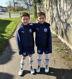 two young boys standing next to each other in soccer uniforms on the side of a road