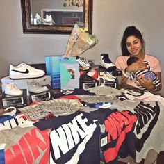 a woman sitting at a table covered in nike shirts and t - shirts, holding a baby