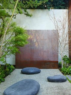 a garden with rocks and trees in front of a white stucco wall that has a red panel behind it