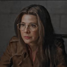 a woman wearing glasses sitting down in front of a brick wall and looking at the camera