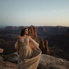 a woman in a long dress standing on top of a cliff with her hands behind her back