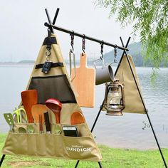 an outdoor kitchen set up with utensils hanging from it's hooks on a tree near the water