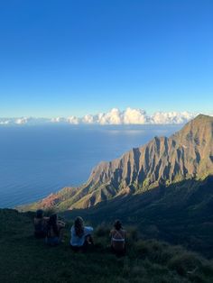 three people sitting on the side of a mountain looking out at the ocean and mountains