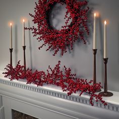 a wreath and candles on a mantel with red berries in the foreground, against a gray wall